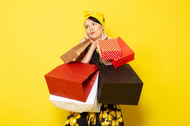 A front view young attractive lady in yellow-black flower designed dress with yellow bandage on head posing holding shopping packages on the yellow